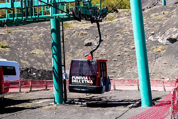 Catania, Sizilien - Italy - 04-22-2025: A red Funivia dellEtna cable car cabin suspended at the base station, surrounded by green support structures and a fenced volcanic landscape.