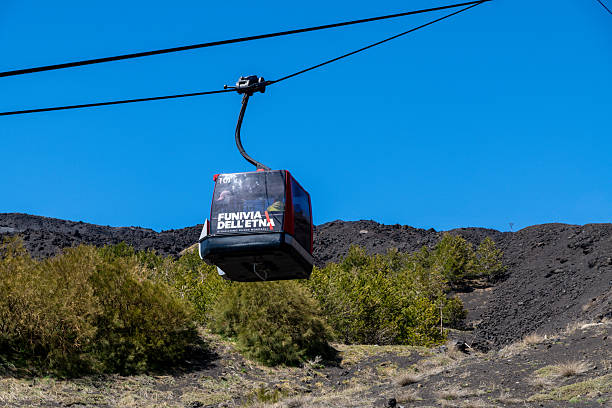 Catania, Sizilien - Italy - 04-22-2025: A red gondola of the Etna cable car travels above green vegetation and volcanic terrain under a clear blue sky on the slopes of Mount Etna.