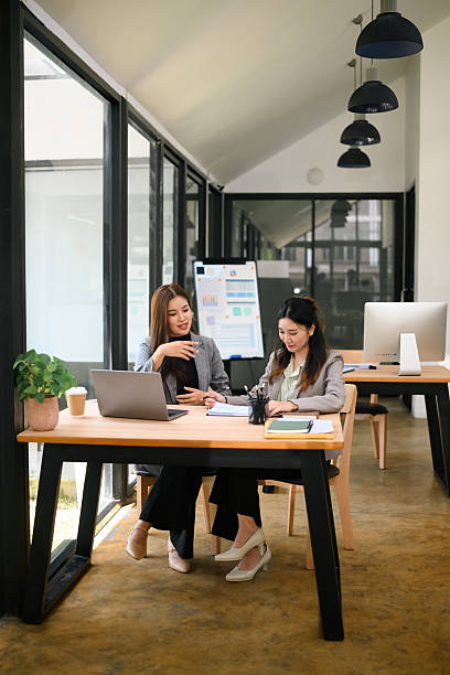 Two confident businesswomen discussing ideas and reviewing documents at a shared desk in a bright modern office