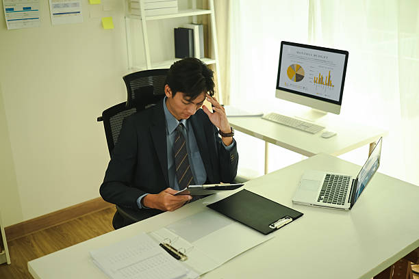 Young businessman in a suit sitting at a desk, appearing stressed while reviewing documents. Office setting with charts on a monitor in the background and a laptop on the table