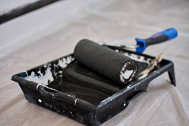 Black paint roller lying in tray filled with black paint on protective film during interior painting process. Preparation for wall painting during home renovation