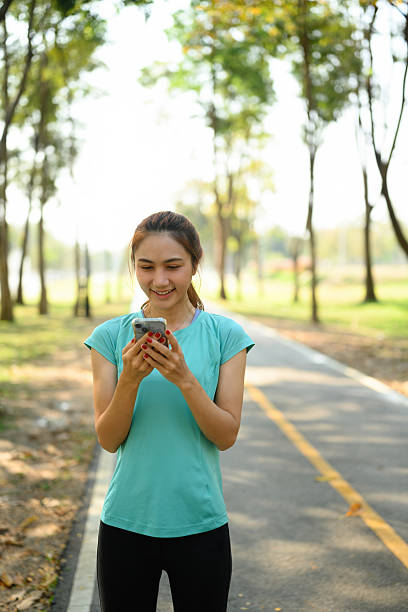 istock Young Asian woman in workout clothes smiles while using her smartphone during a break from exercising in a tree-lined park. Active lifestyle with digital connection in nature 2219329535
