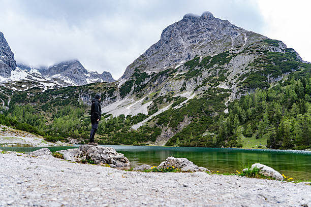 A hiker stands on a rock at the edge of Seebensee, a pristine alpine lake near Ehrwald in Tyrol, Austria. Surrounded by dramatic peaks and reflected greenery, the scene captures the essence of alpine adventure and solitude in the Austrian Alps under a cloudy spring sky.