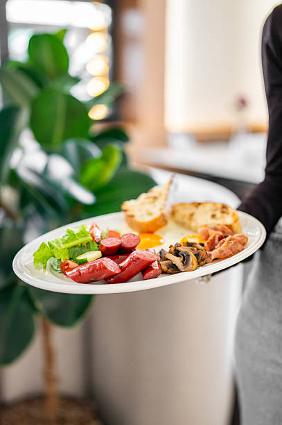 istock A hearty breakfast on a white plate with sausages, bacon, a fried egg, toasted bread, fresh lettuce, cucumber, tomato, and mushrooms. The dish is held by a person in a casual dining setting. 2219336886