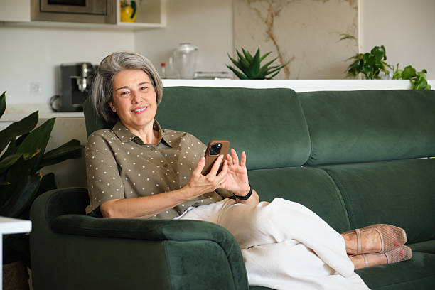 istock Smiling mature woman using smartphone while relaxing on sofa at home 2219337611