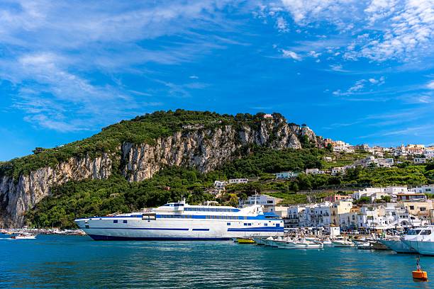 A scenic view of Capri's harbor with a ferry and colorful houses set against a lush green hillside under a vibrant blue sky.
