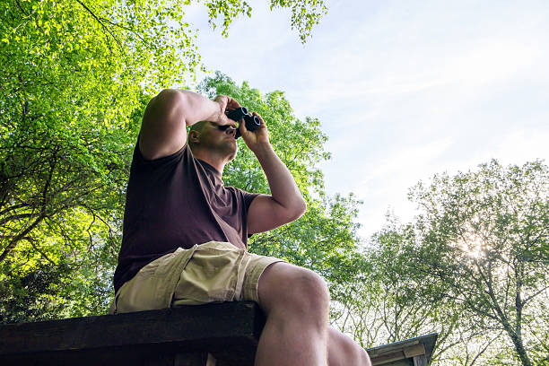 Robust, muscular man sitting on a wooden structure, watching birds through binoculars. Wearing a brown T-shirt and light shorts, surrounded by lush green trees and a partly visible blue sky. Typical birding scene with clear copy space in the sky. A calm, nature-connected atmosphere.