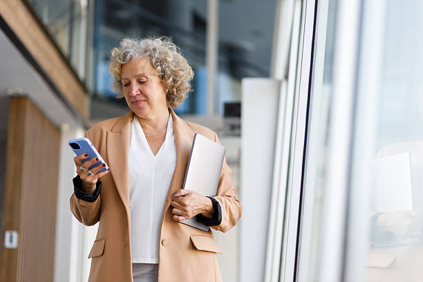 istock Senior businesswoman walking through a modern office building while using her smartphone and carrying a laptop 2219350692