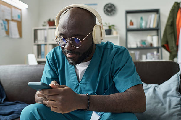African American male nurse wearing headphones, engrossed in checking his smartphone in living room setting filled with bookshelves and notice boards