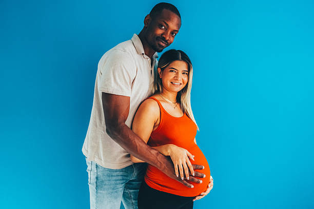Couple stands close together, with the man gently embracing his pregnant partner from behind and both hands resting on her belly. The bright blue background and warm, affectionate pose capture the joy and anticipation of expecting a baby.