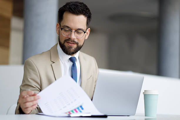 istock Young businessman reviewing financial documents at his desk, using laptop for analysis and strategic decision-making 2219373610
