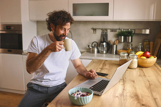 istock Smiling man drinking coffee and using laptop in kitchen 2219375870