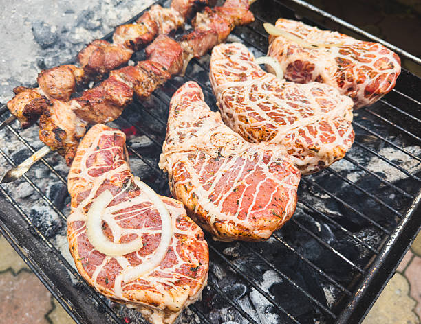 Three pieces of meat are being cooked on a grill. The meat is covered in a white sauce and has onions on top. The grill is on a brick surface