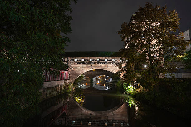 A captivating nighttime view of the historic Weinstadel Bridge in Nuremberg, Germany. The medieval half-timbered architecture is beautifully illuminated, casting warm reflections onto the calm waters of the Pegnitz River