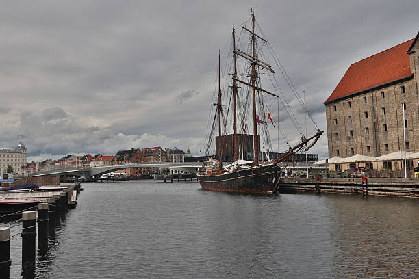 A majestic tall ship gracefully sails through the scenic canals of Copenhagen, Denmark. Its towering masts and billowing sails stand in striking contrast against the historic waterfront, where colorful buildings and charming bridges frame the scene