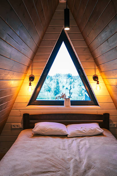 istock Warm bedroom interior in wooden A-frame cabin, bed with pillows, triangular window with forest view, cozy lighting with wall lamps, natural wood decor 2219431976