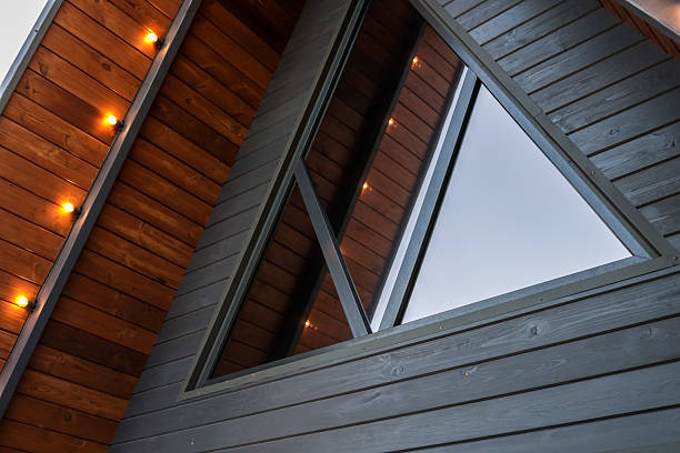 Detail view of modern wooden A-frame cabin facade with large triangular window and warm exterior lights in the evening, minimalist architecture