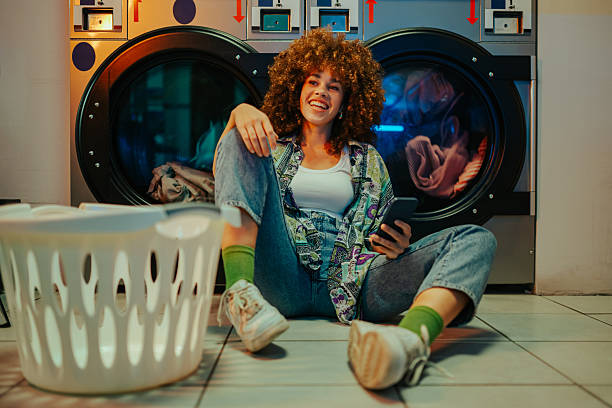 istock Young woman using mobile phone while waiting for laundry in laundromat 2219437441
