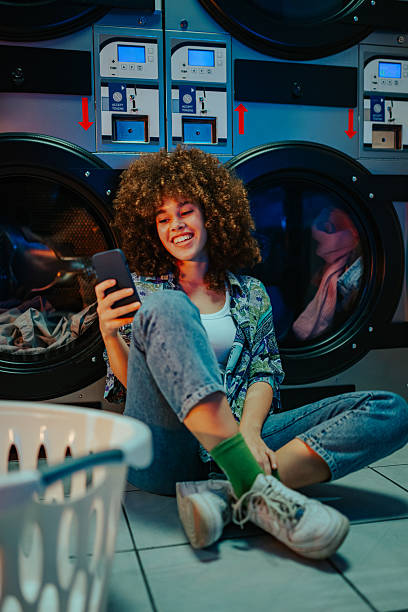 istock Young woman using smartphone while waiting for laundry at laundromat 2219437464
