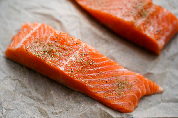 Close-up image of fresh raw salmon fillets seasoned with herbs and spices, placed on parchment paper. The vibrant orange flesh of the fish is highlighted, showing the detailed texture and marbling of the meat. Ideal for illustrating healthy eating, seafood preparation, and gourmet cuisine themes.