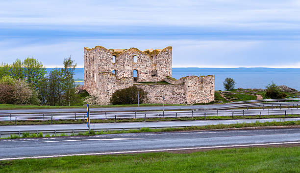 A scenic view of a historic stone ruin beside a highway, with lush greenery and a calm sea in the background under a cloudy sky.