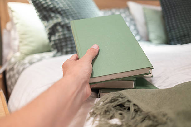 Hand picking up a green book from a stack resting on a bed, surrounded by a cozy blanket and soft pillows, creating a tranquil reading atmosphere in a serene bedroom
