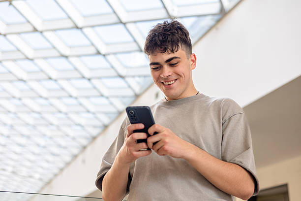 istock Smiling teenage boy using smartphone in modern building. Happy young man texting on mobile phone. Teen boy in gray t-shirt typing on cellphone. young man with smartphone. Copyspace 2219443839