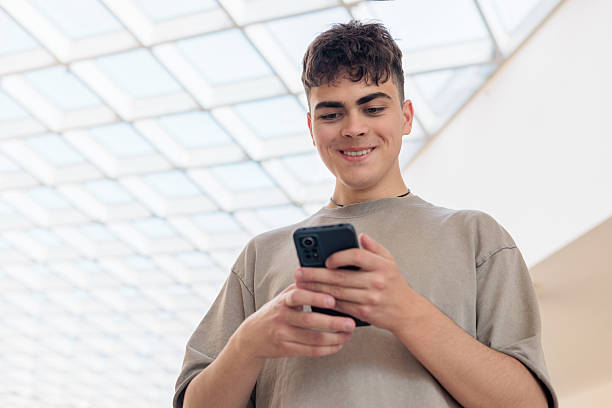 istock Smiling teenage boy using smartphone in modern building. Happy young man texting on mobile phone. Teen boy in gray t-shirt typing on cellphone. young man with smartphone. Copyspace 2219443874