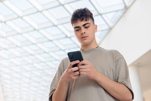 istock Teen boy browsing phone under glass ceiling. Focused male checking mobile device in modern interior. Young adult reading message on smartphone. Concentrated student scrolling through phone. Copyspace 2219443905