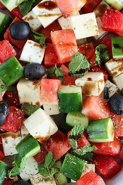 Full frame image of Greek watermelon salad in bowl, with sliced cucumber, red onion, feta cheese cubes, mint leaves and black olives drizzled with balsamic vinegar, blue background, focus on foreground, healthy eating concept