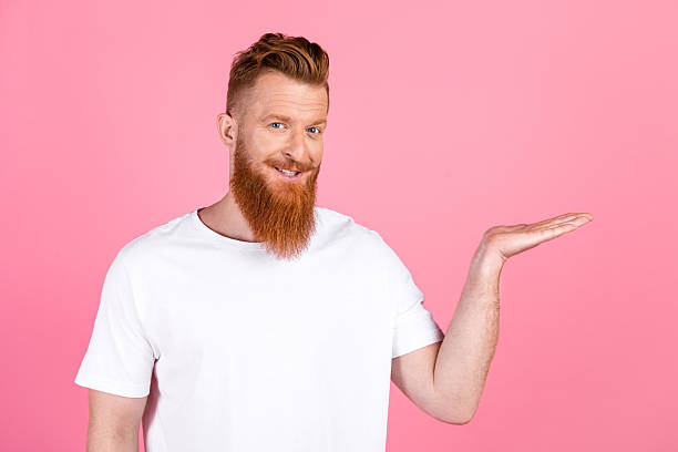 istock Young red haired man gesturing with open palm against pink background while smiling in casual white t-shirt 2219460620