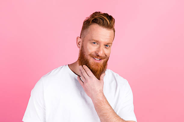 istock A cheerful red-haired man with a beard touching his face on a vibrant pink background wearing a white t-shirt 2219460666