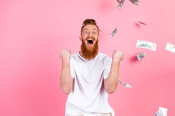 istock Excited young man with red beard celebrating success while surrounded by flying money against pink background 2219460744
