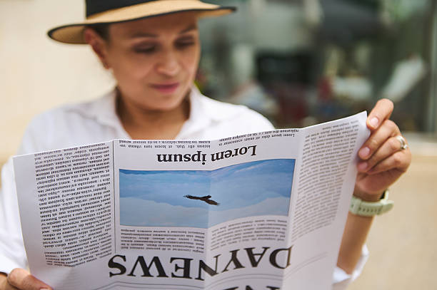 A focused woman wearing a straw hat reading an inverted newspaper outdoors, creating a casual and creative moment with a mix of curiosity and relaxation.