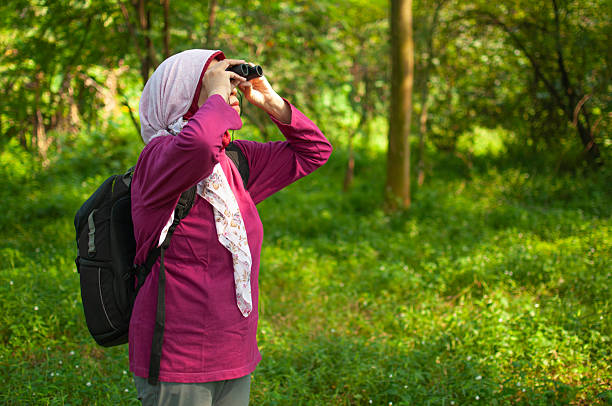 a Middle age Muslim woman holding a binocular and watching bird while walking in the forest