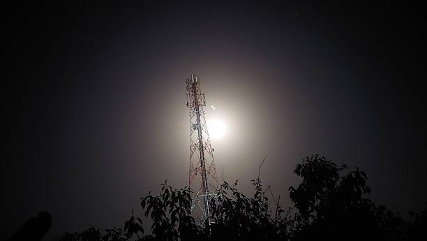 Tall cell tower with multiple antennas stands under a gray sky, with the crescent moon visible in the background. Silhouetted trees and crisscrossing power lines frame the scene, creating a contrast between the technological structure and the natural elements. The evening light casts a serene ambiance over the landscape.