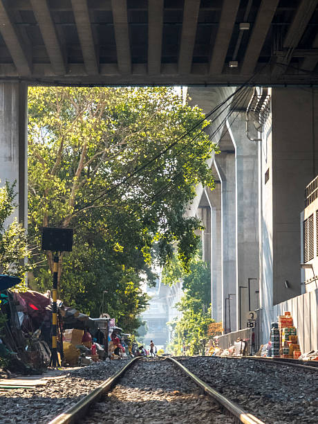 Bangkok,Thailand-April 07 2024-City railway tracks run under the shade of a large concrete overhead rail structure,past trackside shanty dwellings,on a sunny afternoon.