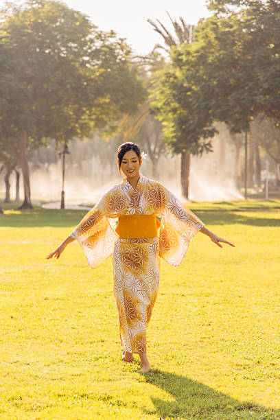 A woman wearing a traditional kimono walks gracefully in a sunlit park during a beautiful day. The serene environment underlines the cultural elegance and relaxed atmosphere of the moment, creating a scene of harmony and beauty.