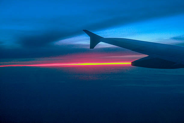 A dramatic view from an airplane window during flight, showing the silhouette of the wing against the backdrop of a bright, colourful sunset on the horizon. The deep blue colour of the sky transitions into bright red and orange hues, evoking a sense of adventure, travel and serenity above the clouds.
