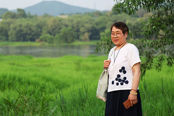 Four elderly women dressed casually in sun hats, standing near a picturesque lake, enjoying their time together, laughing and posing in the warm sunlight surrounded by lush greenery and natural beauty.