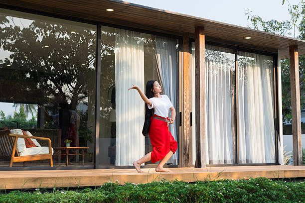 A long-haired Asian teenage girl wearing a white t-shirt and red chong kraben is practicing Thai traditional dancing on her front porch in the warm evening sunlight at home.