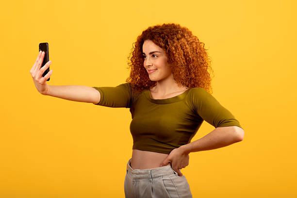 Young woman with curly red hair taking a selfie with her smartphone, smiling and posing on a vibrant yellow background