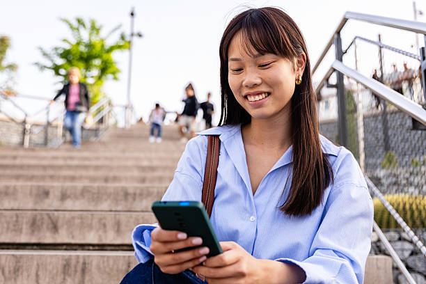 istock Young asian woman using smartphone while sitting on steps outdoors 2219483196