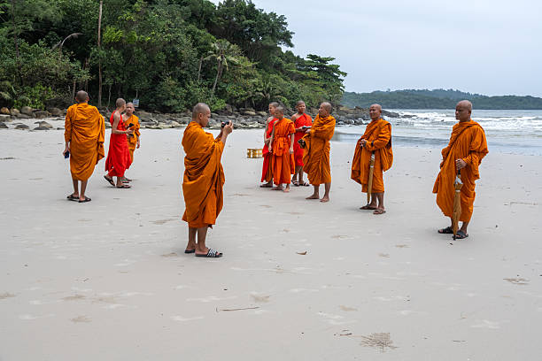 Buddhist monks dressed in vibrant traditional robes are captured enjoying leisure on a scenic beach at Koh Kut, Thailand. The environment exudes tranquility as the monks engage in photography and conversation.