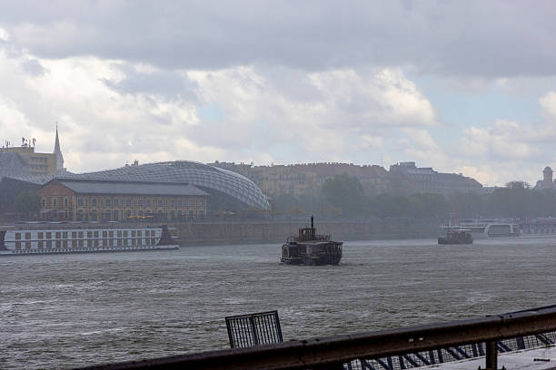 Rainy day view of a river with boats sailing under cloudy skies. The background features a glass-domed building and urban architecture, likely a European city. The image captures the moody, tranquil essence of a cityscape during rainfall