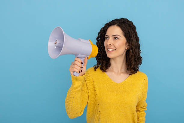 istock Woman holding megaphone, announcing with cheerful expression, glancing sideways against blue backdrop 2219488448