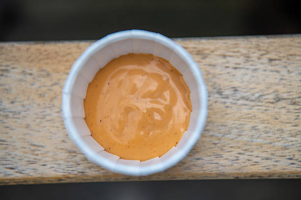 Close-up of a small white paper cup filled with a rich, creamy orange sauce, flecked with seasoning, on a rustic wooden surface. The sauce’s smooth texture and inviting color suggest a flavorful condiment, perfect for food-related themes such as dining, restaurant menus, culinary blogs, or recipe concepts. The warm tones and minimalistic composition create a visually appealing image suitable for stock photography