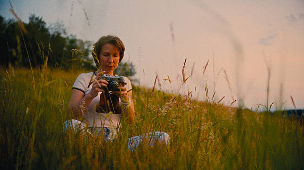 Woman sitting cross-legged,using old-fashioned camera among tall grass in rural field