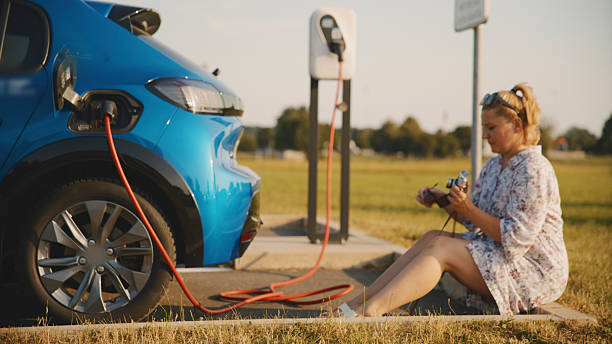 Woman in dress sitting and using retro camera while waiting for electric car to charge in electric vehicle charging station