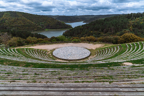 A modern outdoor amphitheater is built into a lush, green hillside overlooking a tranquil river. Inspired by ancient designs, this serene space at the Mimar Sinan Kent Ormani (City Forest) stands empty under a dramatic sky, capturing the harmony between architecture and nature